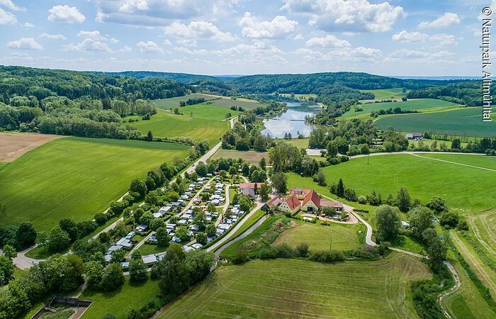 Der Campingplatz Hasenmühle von oben inmitten grüner Wiesen und Bäumen. Hinter dem Platz ist der Hahnenkammsee zu sehen.