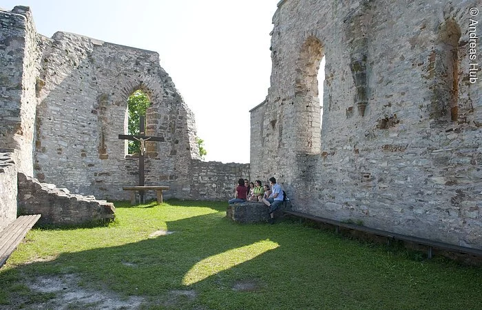 Alte Ruine aus Stein mit Kreuz und davor Menschen, die sitzen.