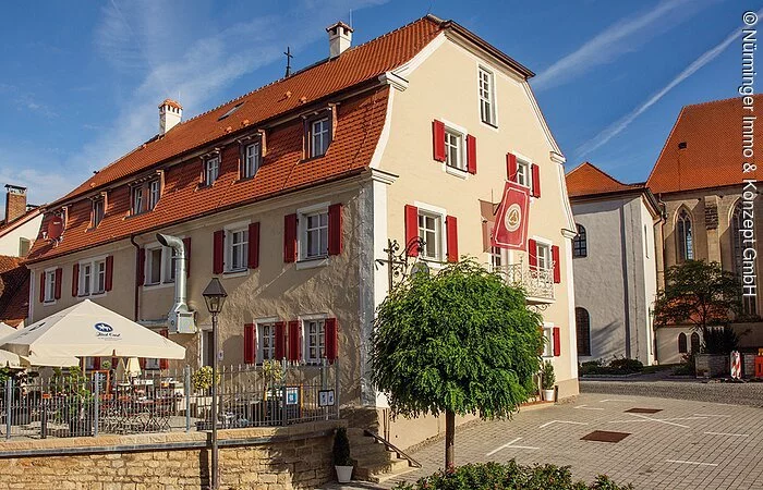 Eine großes beige gestrichenes Gasthaus mit roten Fensterläden. rechts davon ein großer Biergarten mit Sonnenschirmen.