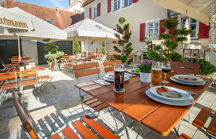 Der Holztisch im Biergarten ist mit einer deftigen Brotzeit gedeckt. Dazu stehen gefüllte Biergläser und ein Weinglas dabei. Im Hintergrund ist ein weißer Sonnenschirm und der restliche Biergarten zu erahnen.