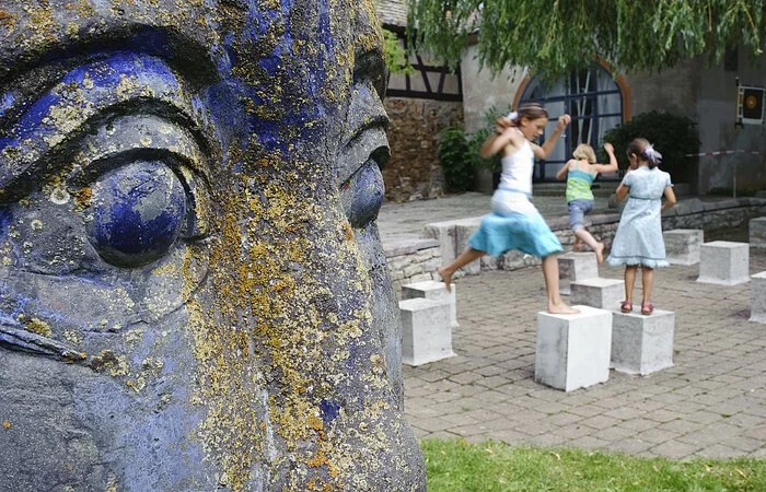 Rechts im Bild die Nahaufnahme des Gesichts einer blauen Statue. Dahinter stehen Steinwürfel in einem Hof. Drei junge Mädchen hüpfen von einem Würfel zum anderen.