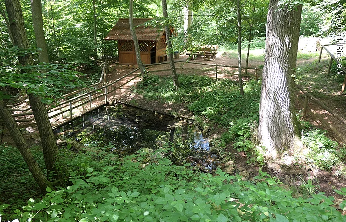 In einer Senke in einem Wald befindet sich ein von Holzzäunen umgebener kleiner Weiher. Im Hintergrund sind eine kleine Holzhütte und Sitzbänke zu sehen.
