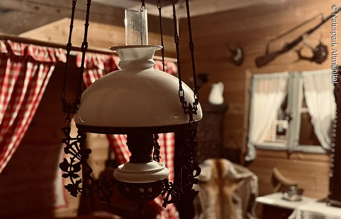 In der Mitte des Bildes hängt eine Öllampe von der Decke über einem Tisch. Im Hintergrund ist eine Fensterbank mit roten Vorhängen zu sehen.