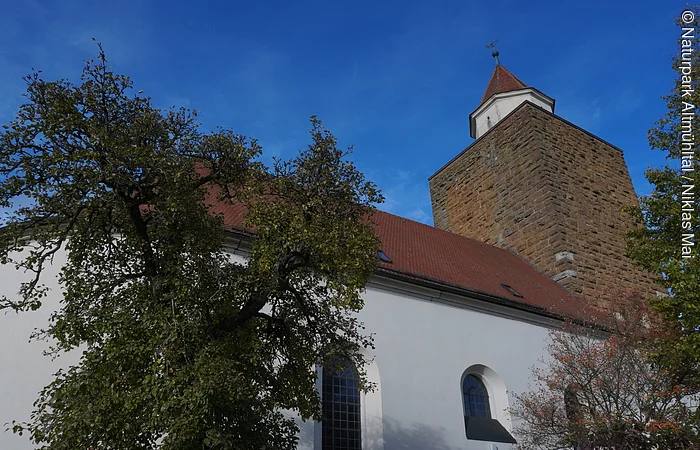 Kirche mit weißer Fassade, rotem Dach und steinernem Turm vor blauem Himmel und Bäumen im Vordergrund.