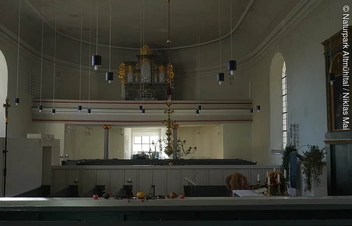 Innenraum einer Kirche mit Orgel, Kronleuchter, Kerzen und Fenstern an den Seitenwänden.