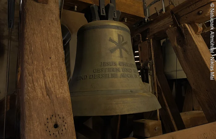 Große bronzene Kirchenglocke mit christlichem Symbol und Schriftzug, umgeben von Holzbalken im Glockenturm.